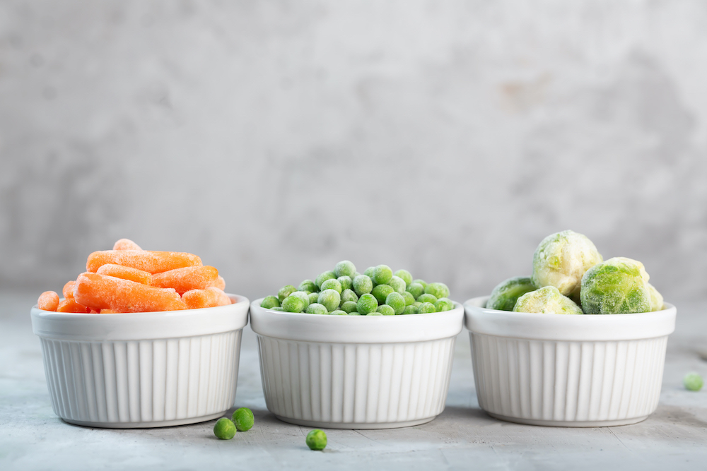 Frozen vegetables such as green peas, brussels sprouts and baby carrot in the white bowls on the concrete gray background